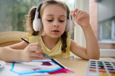 Close-up portrait of adorable European preschool child, cute baby girl in audio headset, painting picture with brush and watercolor. Creativity courses for children, talented little artists, painters clipart