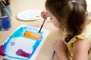 Focus on unfinished picture on table, and blurred little girl painting with paintbrush and watercolors during art class. Overhead view
