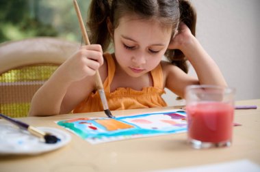 Concentrated Caucasian smart little preschooler girl, painting picture with watercolors and paintbrush. Drawing. Art class. Painting