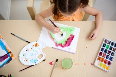Top view of a Caucasian little girl, talented child drawing picture with paintbrush and watercolors. Painting art class for kids clipart