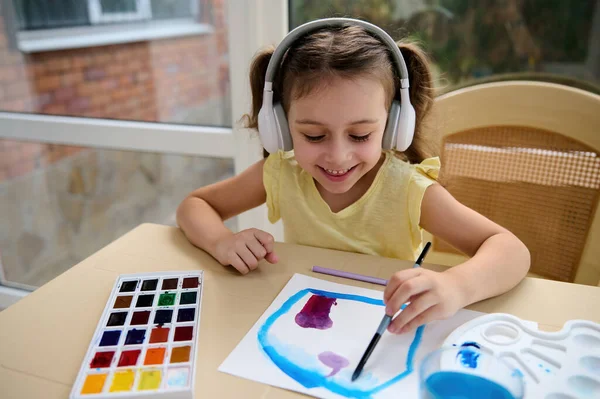 Happy child, adorable European little girl of preschool listens to the music in wireless headphones, learns watercolour painting. Back to school. Homeschooling. Kids development and art education