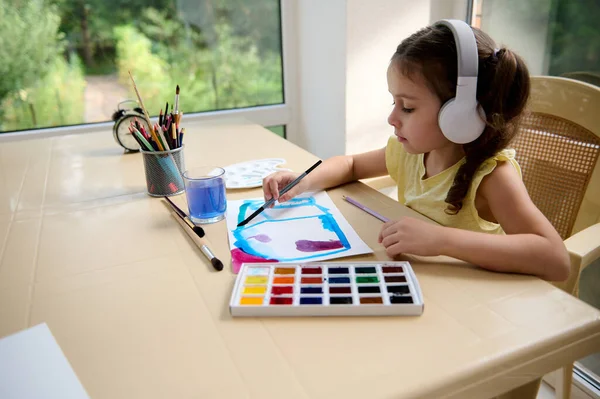 Relaxed beautiful talented Caucasian little girl of preschool, wearing wireless headphones, painting picture with watercolors. Drawing, artistic creativity concept. Education, childrens entertainment