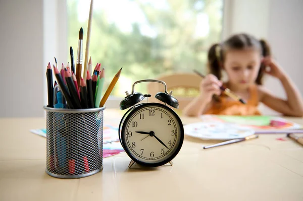 Focus on black alarm clock and pencil holder on the blurred background of little girl of preschool age, sitting at table and drawing picture with watercolors. Painting. Art class and courses for kids