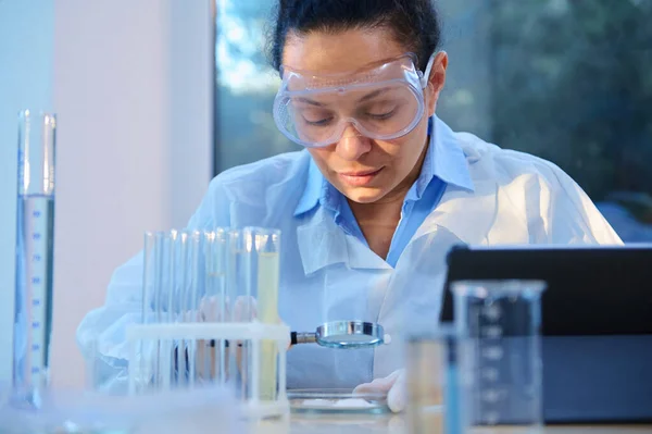 Female scientist using a magnifying glass examines biological material in Petri dish, sitting at a table with lab glassware and a digital tablet, conducting clinical research in a medical laboratory