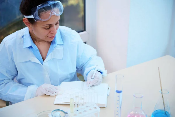 Portrait of a female scientist in white lab coat and protective goggles writing on a notepad the results of conduct scientific experiment, sitting at a table with lab glassware in chemical laboratory