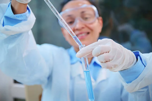 Selective focus on a glass graduated laboratory pipette in the hands of a smiling female clinical research laboratory worker, titrating a chemical reagent into a test tube while conducting experiment