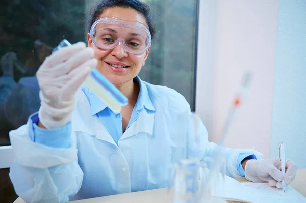 Pleasant African American woman, lab technician, scientist, pharmaceutical chemist holding test tubes with chemical substances, testing the scientific experiment in a clinical research laboratory.