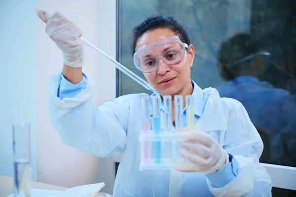 Confident African American pharmacologist, medical biologist, laboratory assistant using lab pipette, pipetting liquid substance into test tubes while doing experiment in clinical research biolab