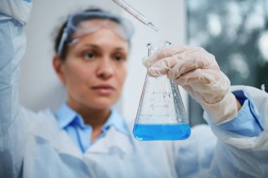 Selective focus on flat-bottomed flask with liquid substance and blurred charming woman scientist, medical biologist using a glass graduated laboratory pipette, dripping few reagent into blue solution
