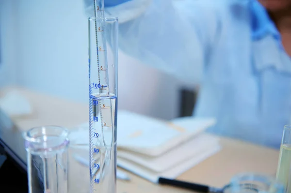 Selective focus on a glass pipette in a graduated cylinder over blurred scientist pharmacologist, making clinical study, working on new vaccine or drug, in a medical science laboratory. Close-up.