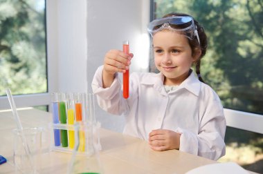 Pretty child chemist, scientist, Caucasian preschooler little girl wearing safety goggles and lab coat doing chemistry experiments, watching the fascinating chemical reaction in the test tube.