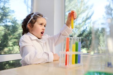 Delighted European primary schoolgirl, in white lab coat and goggles watches a fascinating chemical reaction, between multi-colored chemicals and reagents taking place in test tube at chemistry course