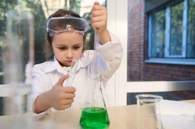 Adorable little girl filling a pipette with green liquid from flat bottomed flask, dropping the reagents into a test tube, while doing a chemistry experiment in chemical scientific school laboratory