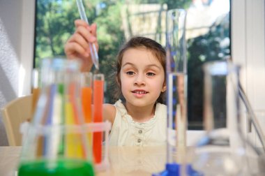 Inspired Caucasian little schoolgirl using glass pipette, makes lab experiment on the Chemistry lesson. Laboratory glassware with chemicals and reagents on the blurred foreground.