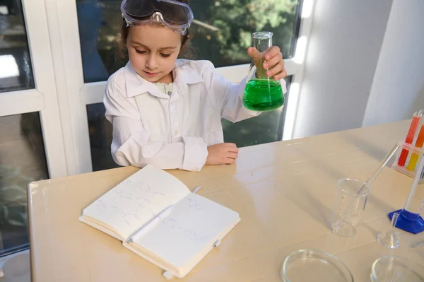 Charming little girl in lab wear, shaking a flask with green solution while reading the chemical formula in chemistry textbook in the school scientific laboratory. Back to school in new academic year