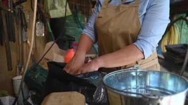 Close-up hands of florist, gardener in beige apron standing at a wooden table with lots of garden tools, sorting fertilized soil with her hands in the summer cottage workshop. Gardening. Horticulture