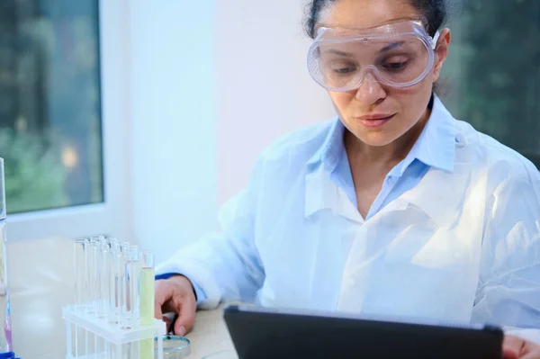 Portrait of a multi-ethnic woman, scientist in protective goggles and white lab coat, using on a digital tablet, analyzing experimental results in a medical laboratory. Pharmacology. Clinical research