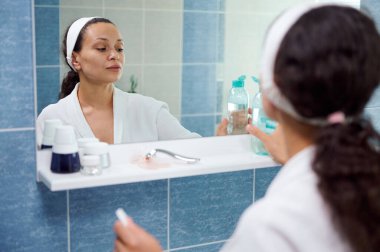 Charming Latin American middle-aged woman in white bathrobe, standing in front of a bathroom mirror and holding a face cleanser, to remove make-up and clean her face. Beauty treatment and skin care