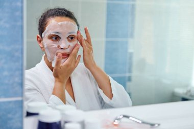 Charming dark-haired multi-ethnic woman in white bathrobe, looking at mirror in bathroom while applying moisturizing smoothing tissue mask on her face, taking care of the beauty and health of her skin