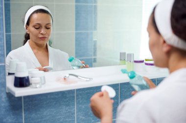 Reflection in bathroom mirror of a dark-haired African American young pretty woman, wearing white bathrobe, using a cotton pad and a face cleanser to remove make-up. Healthy skin is a daily commitment