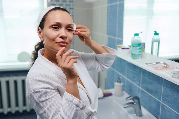 Beautiful multi-ethnic woman in white waffle bathrobe, using a cotton pad, cleans her face with a face cleanser and removes make-up, standing in front of a mirror in the home bathroom