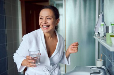 Happy charming multi-ethnic woman in white bathrobe, stands in the bathroom, smiling a cheerful toothy smile, looking away, holding glass of water and taking medicine according to medical prescription