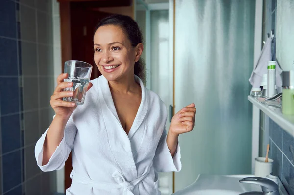 Portrait of a happy young Latin American young woman taking daily dose of complex healthcare skin, hair and nails omega vitamins and drinking glass of fresh pure water. Immunity improvement concept.