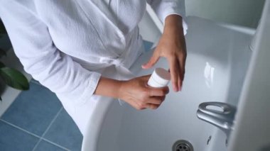 Steadicam shot: Pretty woman in white bathrobe, stands at a washbasin in bathroom, opens a container with pills and takes medication with daily dose of vitamins, minerals and nutritional supplements