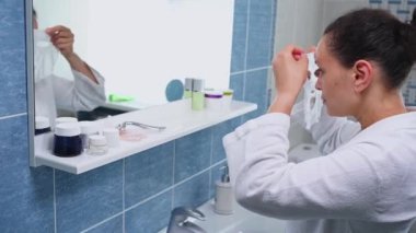 Beautiful multi-ethnic middle-aged woman in bathrobe, stands in front of bathroom mirror, applies a smoothing moisturizing textile facial mask on her face. Home spa, beauty treatment. Selective focus
