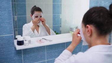Reflection in bathroom mirror of a beautiful dark-haired multi-ethnic woman, in white bathrobe applying a smoothing moisturizing tissue face mask. Skin care. Home spa. Anti-aging beauty procedures