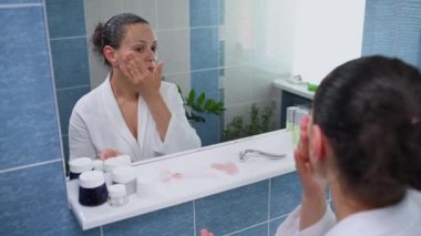 Attractive woman in a white bathrobe, looking at her reflection in bathroom mirror, applying hydrating cream or moisturizing serum and using a pink quartz roller massager performing a lifting massage