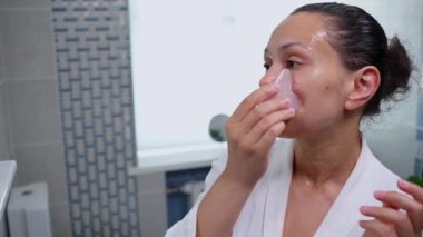 Middle aged pretty woman in bathrobe applies a moisturizing serum to her face and using Gua Sha rose quartz scraper, performs smoothing rejuvenation massage. Alternative rejuvenating physical therapy