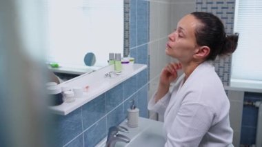 Beautiful middle-aged woman wearing a white bathrobe, standing in front of a bathroom mirror, performing an anti-wrinkle massage on her neck with a Gua Sha rose quartz scraper. Anti-aging treatment