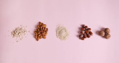 Flat lay stacks of organic raw vegan superfood: sesame seeds, almond nuts, oat-flakes, walnut and hazelnut scattered on pink background, copy space. Horizontal studio shot. Still life. Selective focus