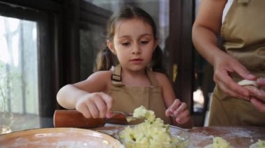 Adorable Caucasian little girl in chef's apron, stuffs mashed potatoes into rolled out pastry molds while her mom hands-on dumplings, enjoying cooking dinner together in the summer country kitchen