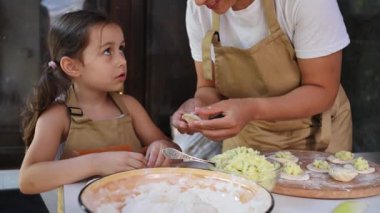 Loving mother in beige chef's apron, stands by a table with dough, teaches her adorable little daughter how to make dumplings, having fun together in the country kitchen. Vareniki Pelmeni Ravioli.