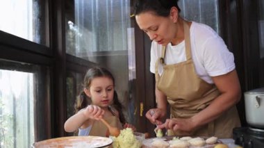 Beautiful child, a little girl in a beige chef's apron, standing next to her mother in the kitchen of a summer cottage, repeating the molding of dumplings, enjoying cooking Ukrainian varenyky together