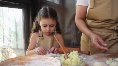 Adorable European child girl with two ponytails wearing chef apron, concentrated on molding dumplings stuffed with mashed potatoes, helping her mother in the kitchen. Mom and daughter cooking together