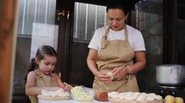 Cheerful mom and daughter wearing chef's aprons, sculpt dumplings, then give hi five and cutely smile at camera, standing with crossed arms in the country kitchen. Woman and little girl enjoy cooking