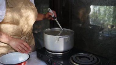 Close-up of a woman housewife in beige chef's apron, standing by countertop and stirring homemade dumplings in the pot with boiling water, preparing Ukrainian national dish varenyky in rustic kitchen