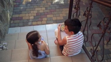 View from the back of children in summer wear, teenage boy and preschool girl, brother and sister enjoy a delicious lunch together, eat homemade dumplings, sit on the stairs of the country house porch
