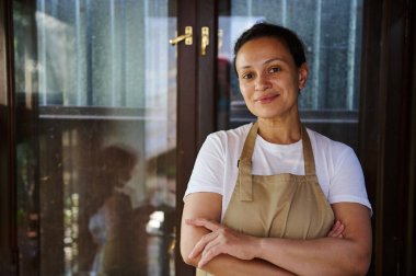 Confident portrait of a multi-ethnic pretty woman, pleasant housewife cook wearing white t-shirt and chef apron, smiles and poses with crossed arms looking at camera standing against rustic background