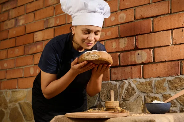 Latin American woman baker confectioner in black chefs apron and white cap, stands against brick wall of artisanal family bakery, and sniffs a loaf of traditional fresh baked whole grain wheat bread