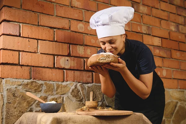Charming Latin American woman, a baker confectioner in chef\'s hat, holds and sniffs a loaf of warm traditional homemade whole grain sourdough wheat bread, freshly baked in the artisanal family bakery