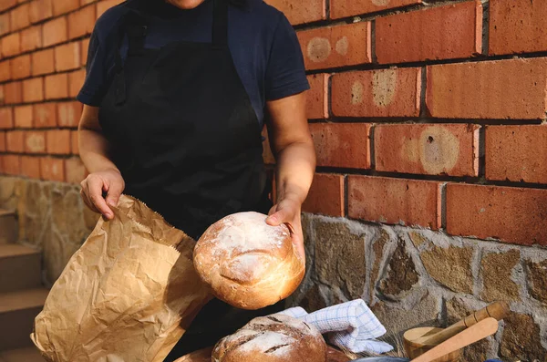 Cropped view of a baker dressed in black, standing against brick wall, holding a loaf of traditional whole grain bread and putting it into a recyclable eco paper bag for sale in artisanal bakery store