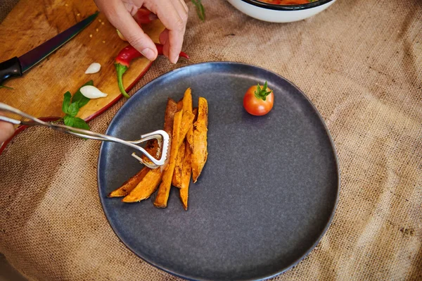 View from above of a chefs hand serving plate, putting sweet potato fries on a dark ceramic plate. Copy advertising space. Web banner. Menu for restaurants. Cooking master class. Healthy vegan food