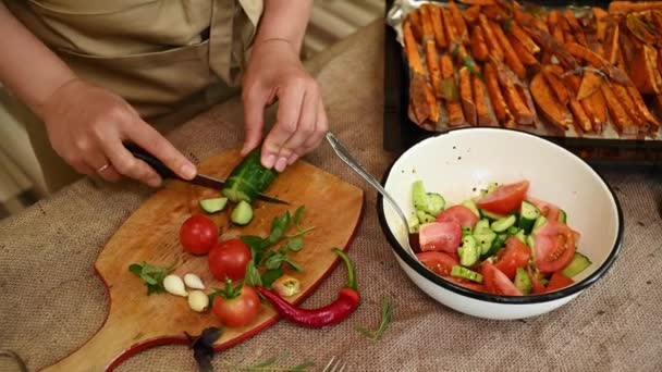 从上面看一个素食主义者的手 一个穿着大厨围裙的家庭主妇 把新鲜黄瓜和西红柿切成薄片 放在木制的切菜板上做沙拉 健康饮食 — 图库视频影像