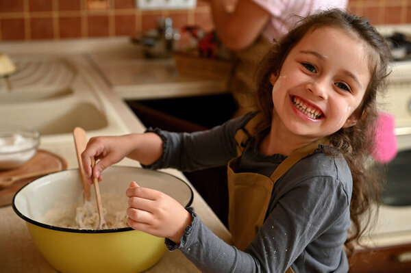 Portrait of a Caucasian cheerful and mischievous little confectioner, an adorable baby girl in a chefs apron, smiling cheerfully, looking at the camera and kneading dough to bake a festive cherry pie