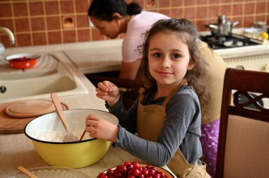 Güzel beyaz çocuk, sevimli küçük kız annesine mutfakta yardım ediyor, yemek yapmayı öğreniyor, hamur yoğuruyor, vişneli turta hazırlıyor, kameraya gülümsüyor.