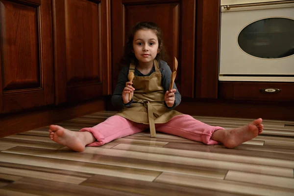 Baby girl, pretty little baker confectioner wearing beige chef apron and pink pants, playing with wooden spoons while sitting barefoot on the kitchen floor, leaning against cupboard with mounted oven
